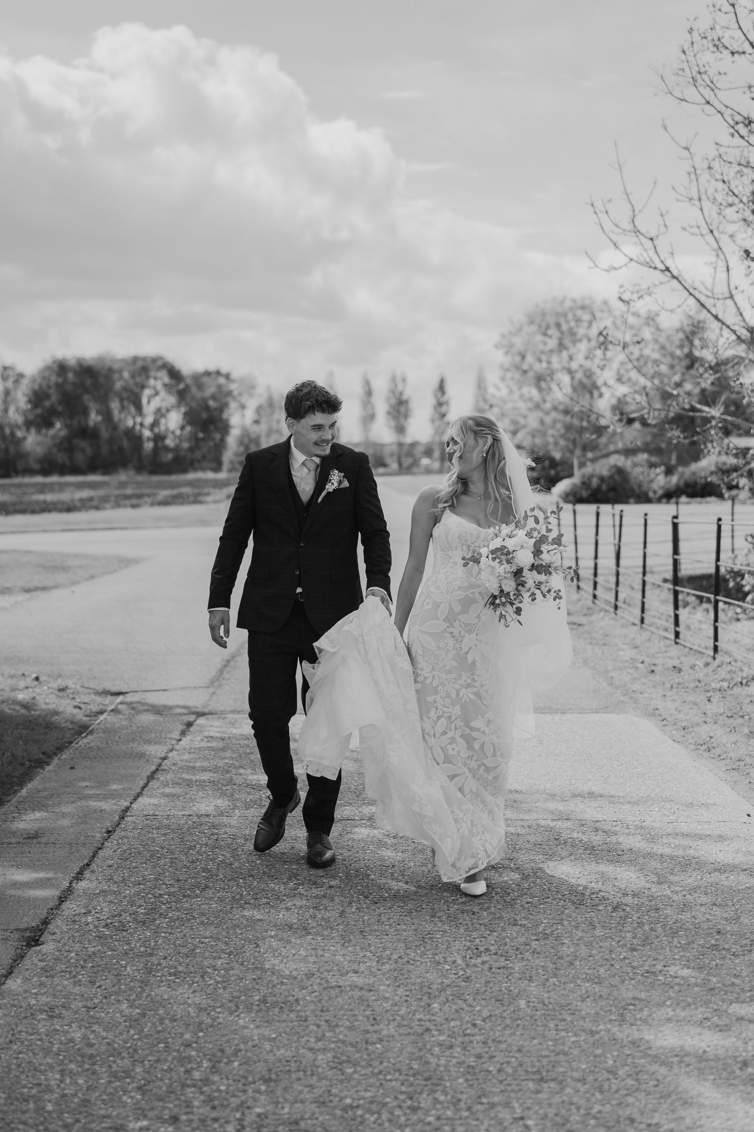 Black and white photo of a bride and groom walking hand in hand on a path, smiling at each other on their wedding day.