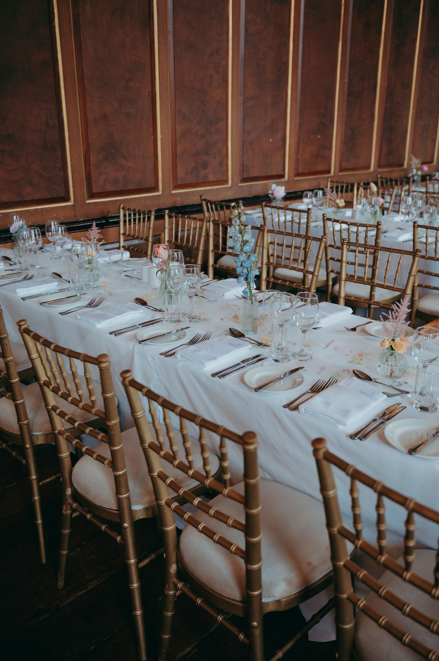 Elegant wedding reception hall with gold chiavari chairs and long white linen tables set with floral centerpieces and gold cutlery.
