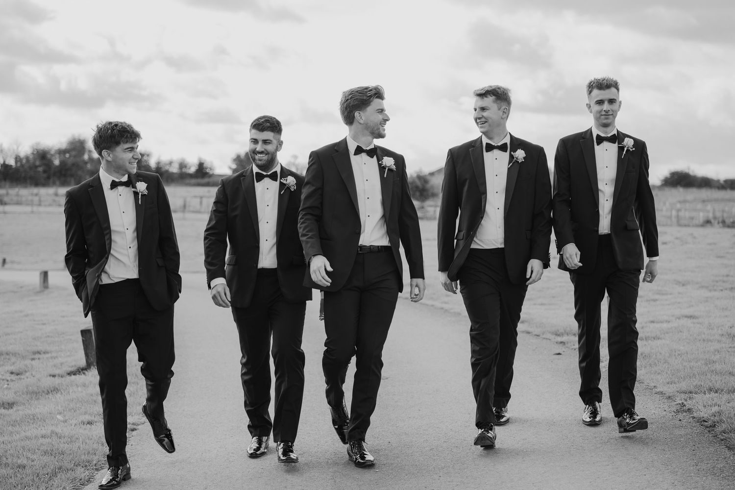 Five groomsmen in black tuxedos and bow ties walk confidently together outdoors in a classic black and white wedding photo.