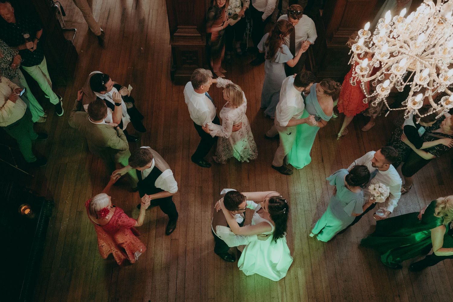 Aerial view of couples dancing at a wedding reception on a wooden dance floor under a chandelier.