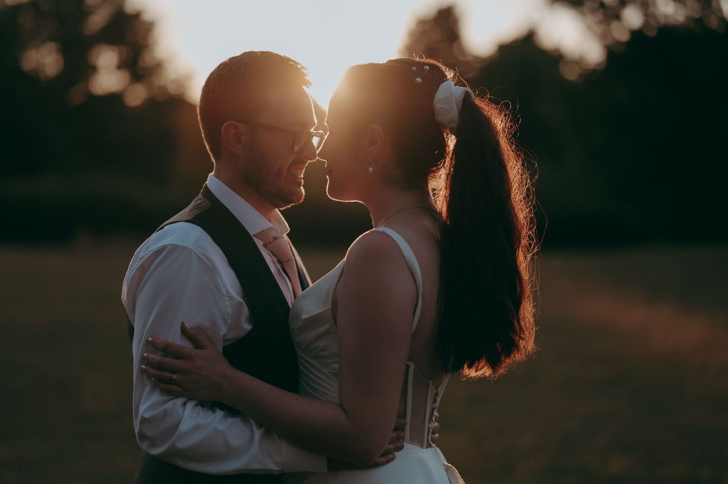A couple shares a romantic sunset kiss outdoors, bride in white gown with floral hair accessory, groom in vest.