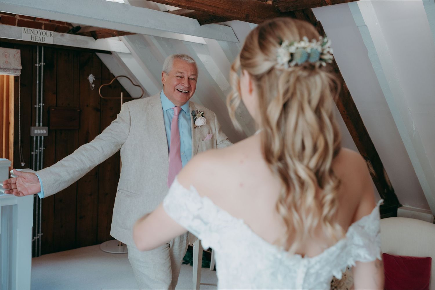 Bride in off-shoulder lace gown shares first look moment with smiling older man in beige suit at rustic venue.