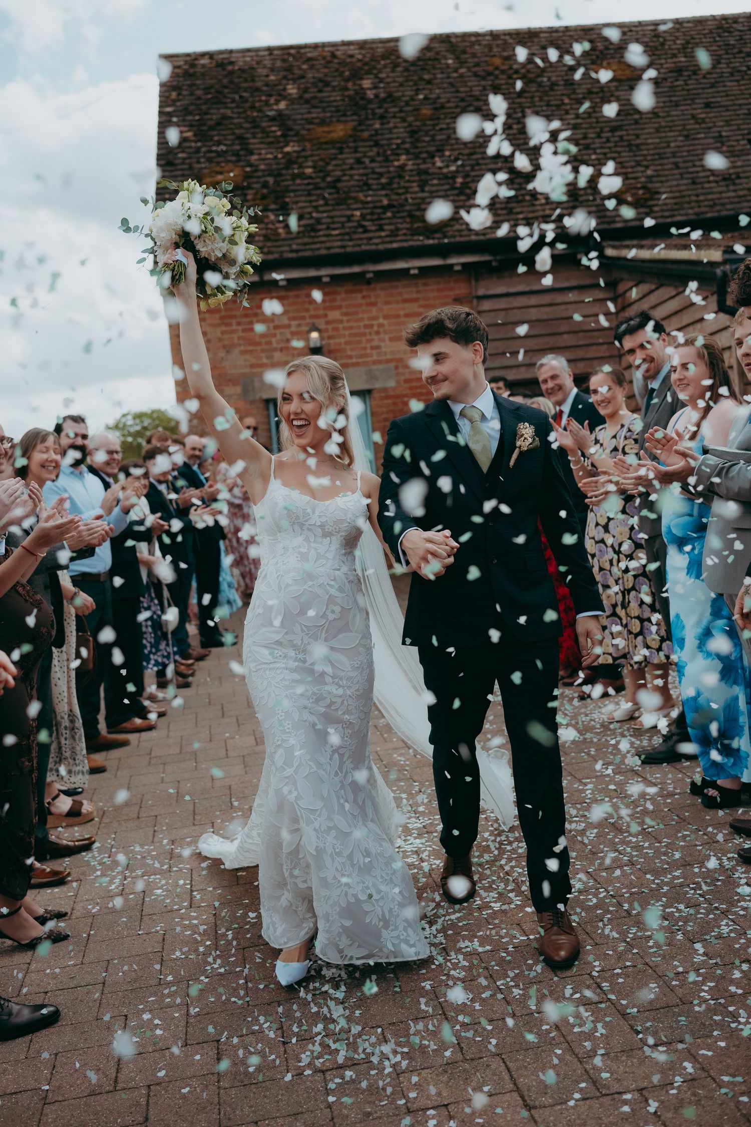 Bride and groom walk through confetti shower, guests cheering outside rustic barn wedding venue on sunny day.