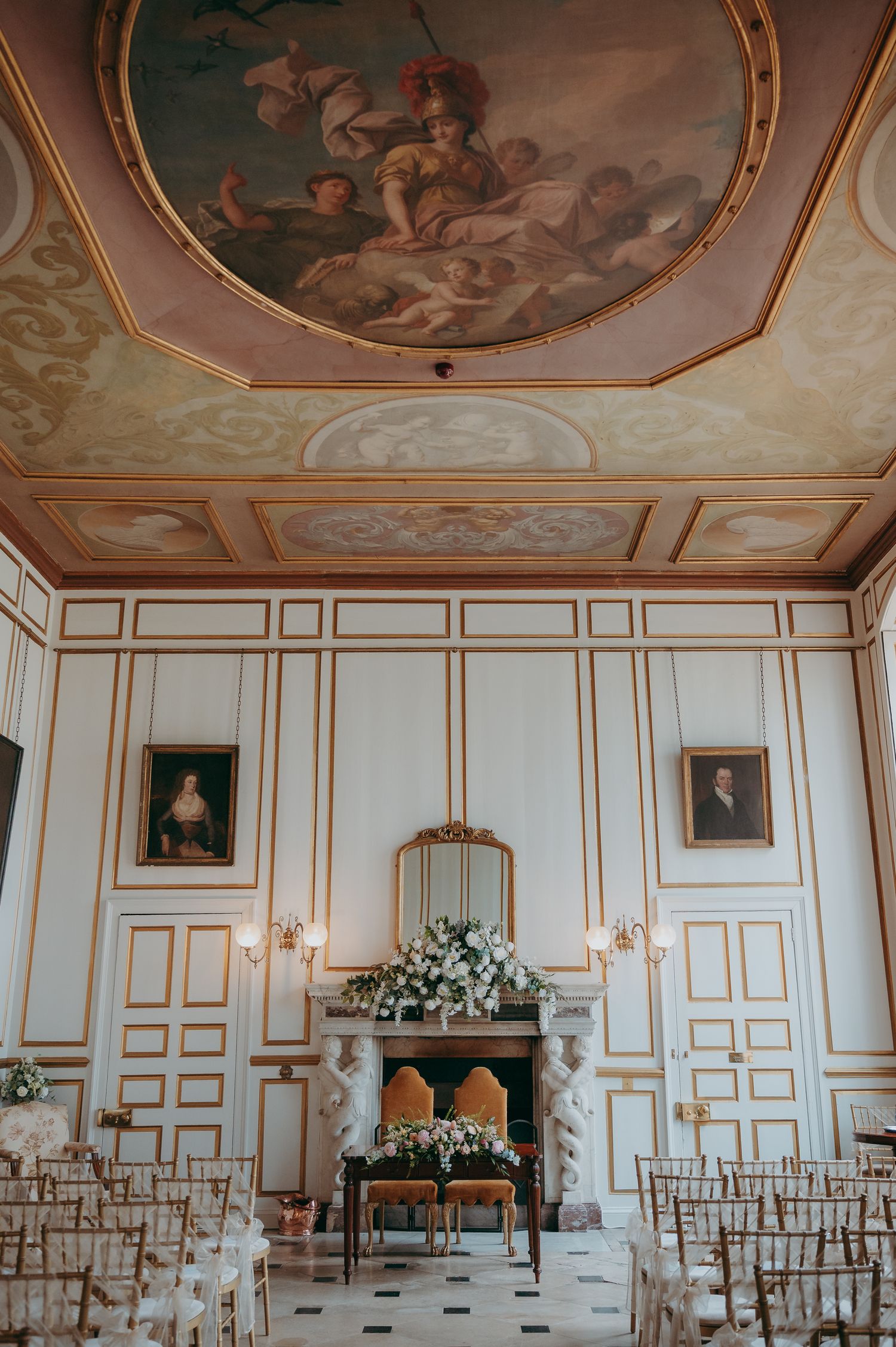 Elegant wedding ceremony room with ornate ceiling mural, gold-trimmed white walls, floral fireplace arrangement, and rows of chairs.