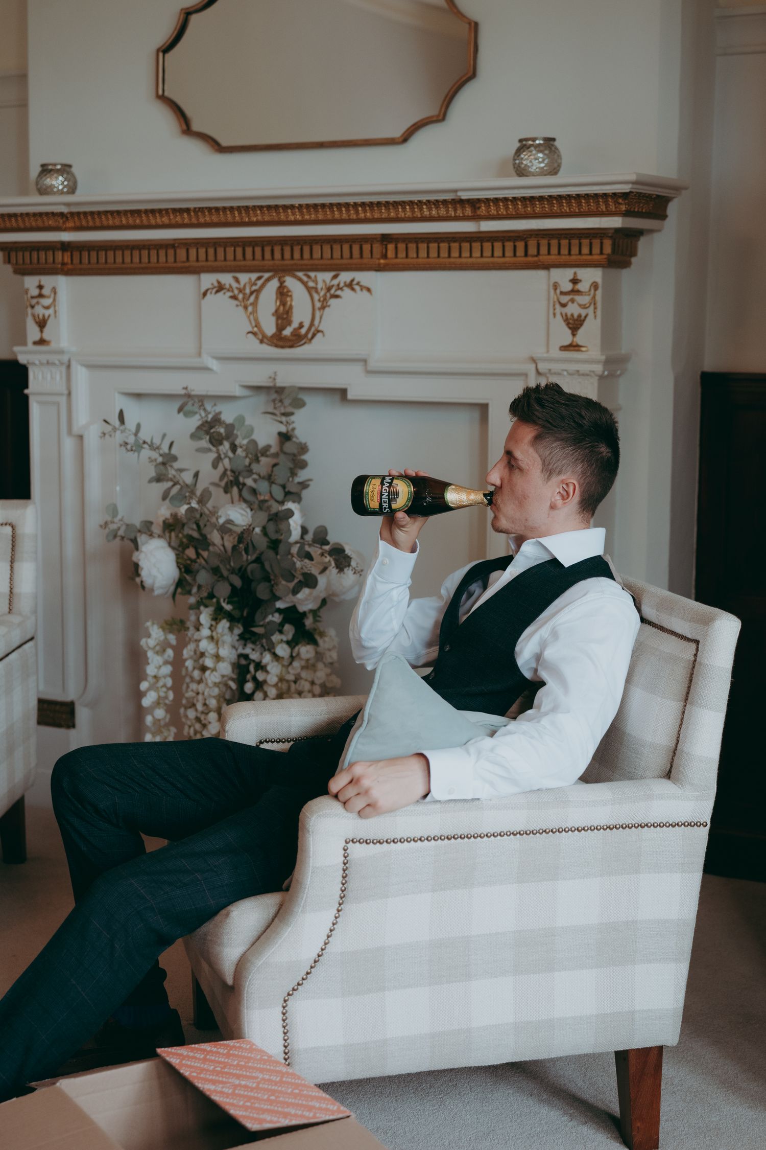 A well-dressed man in a vest sips champagne while relaxing in an armchair near an elegant white fireplace.