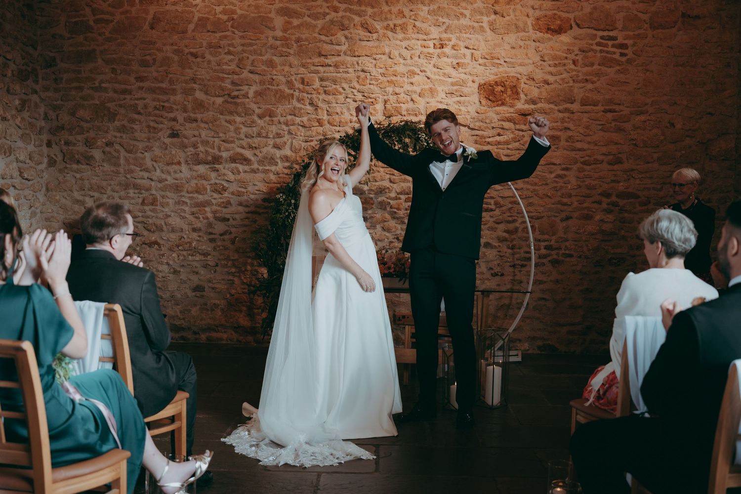 Joyful newlyweds raise hands during rustic barn wedding ceremony with seated guests and brick wall backdrop.