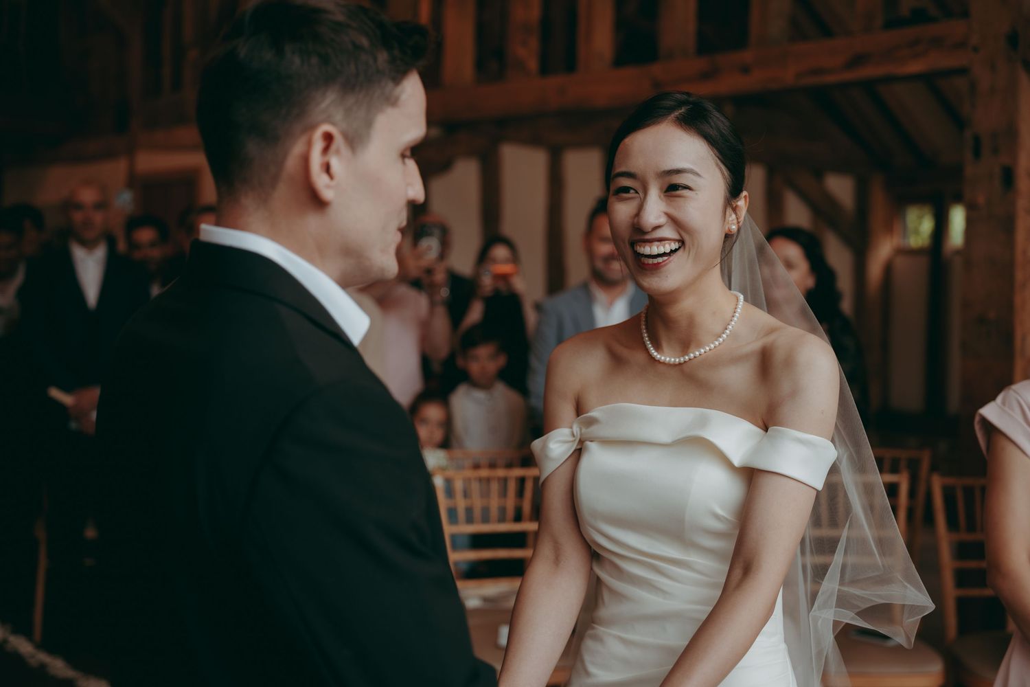 Bride in off-shoulder white gown laughing joyfully with groom during wedding ceremony in rustic barn venue.