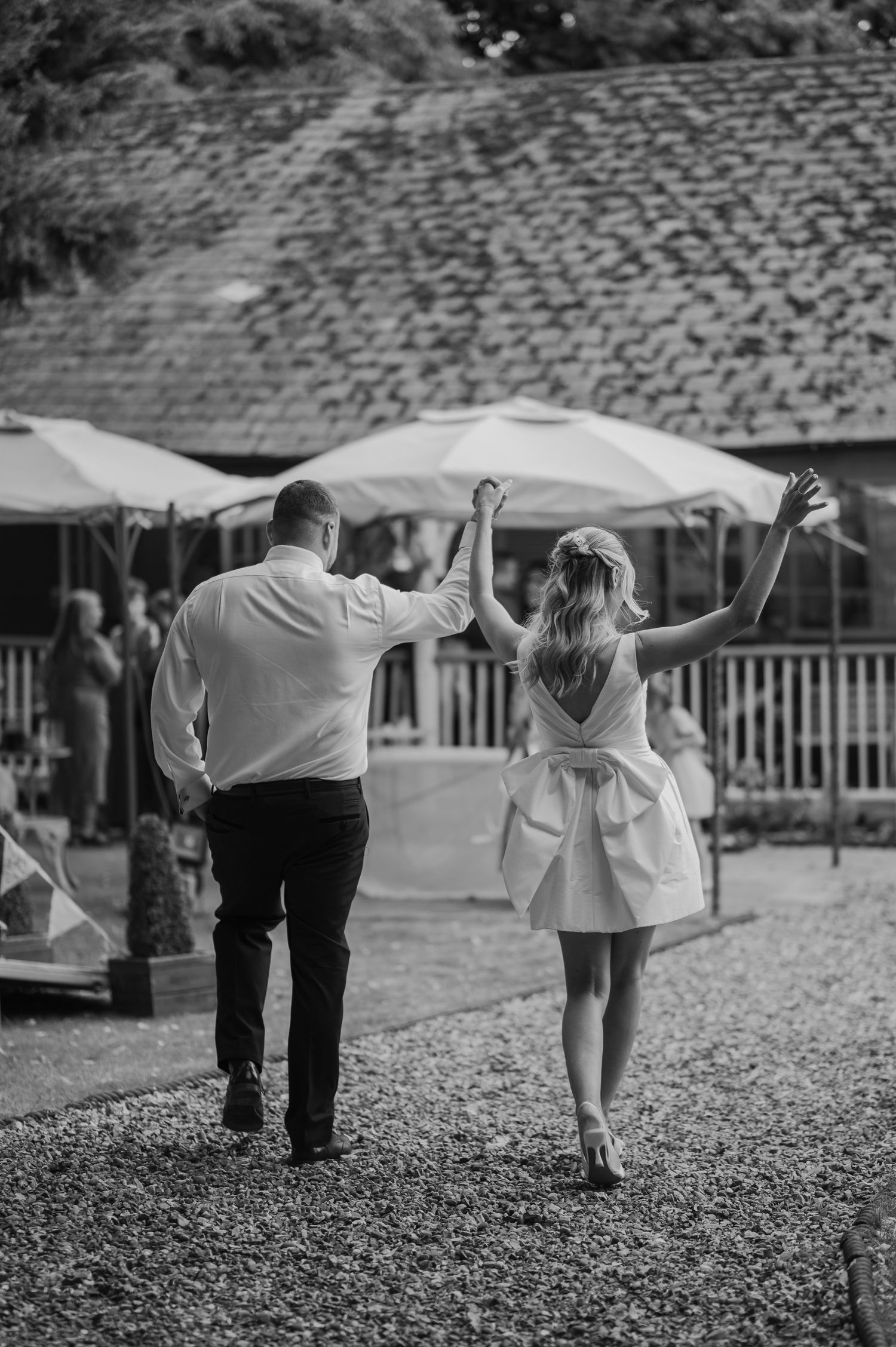 Black and white photo of a bride and groom holding hands, walking toward a rustic venue with umbrellas.