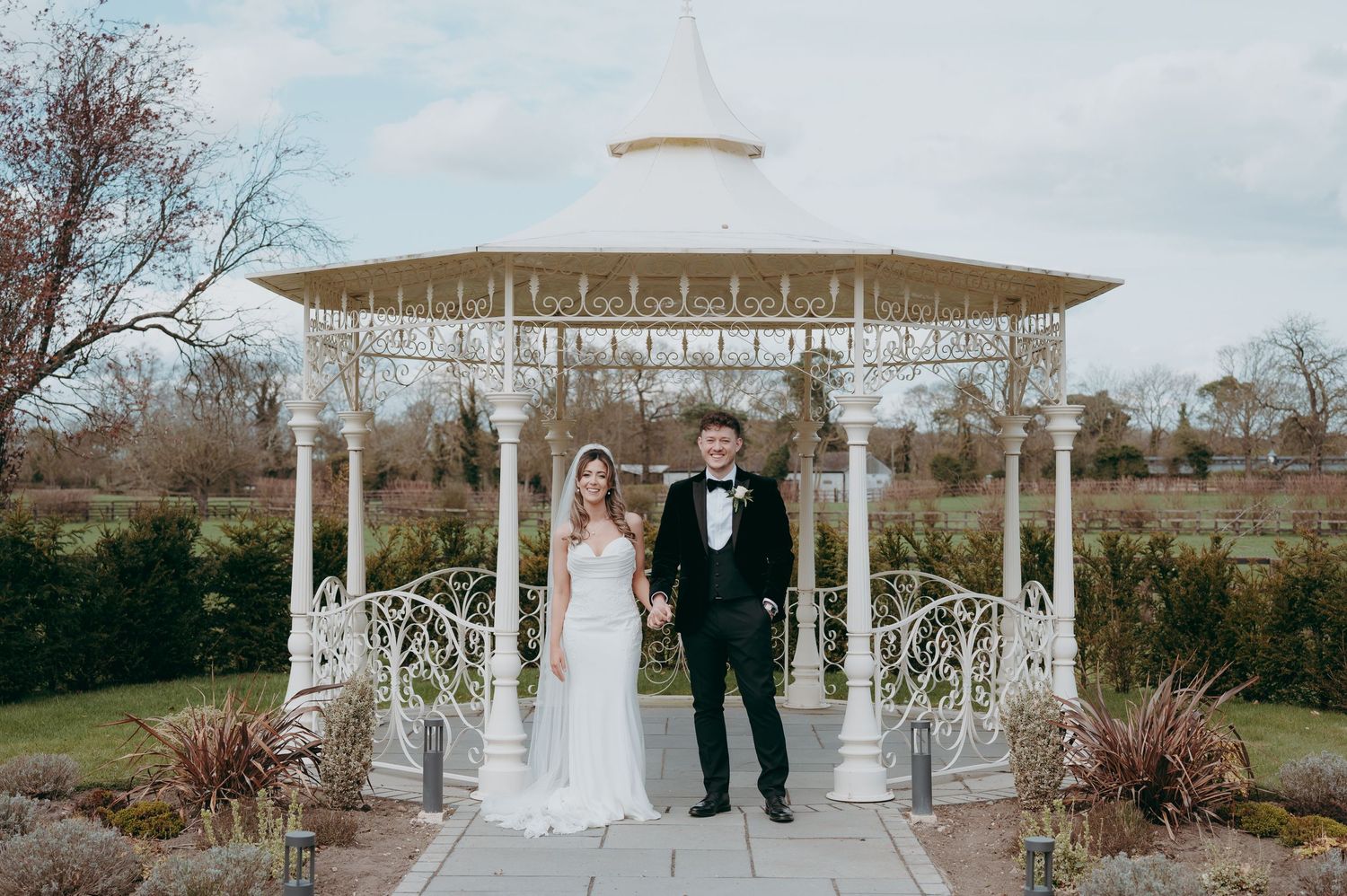 Bride and groom posing together in front of a white ornate gazebo in a lush garden wedding venue.