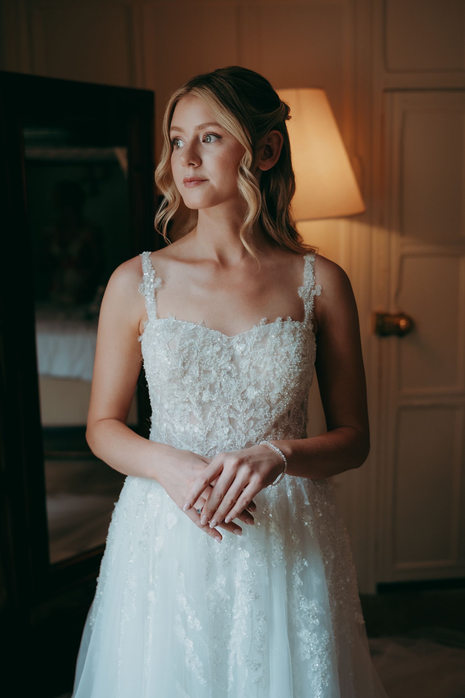 A bride in a floral lace A-line wedding gown stands in a warmly lit room, gazing thoughtfully to the side.