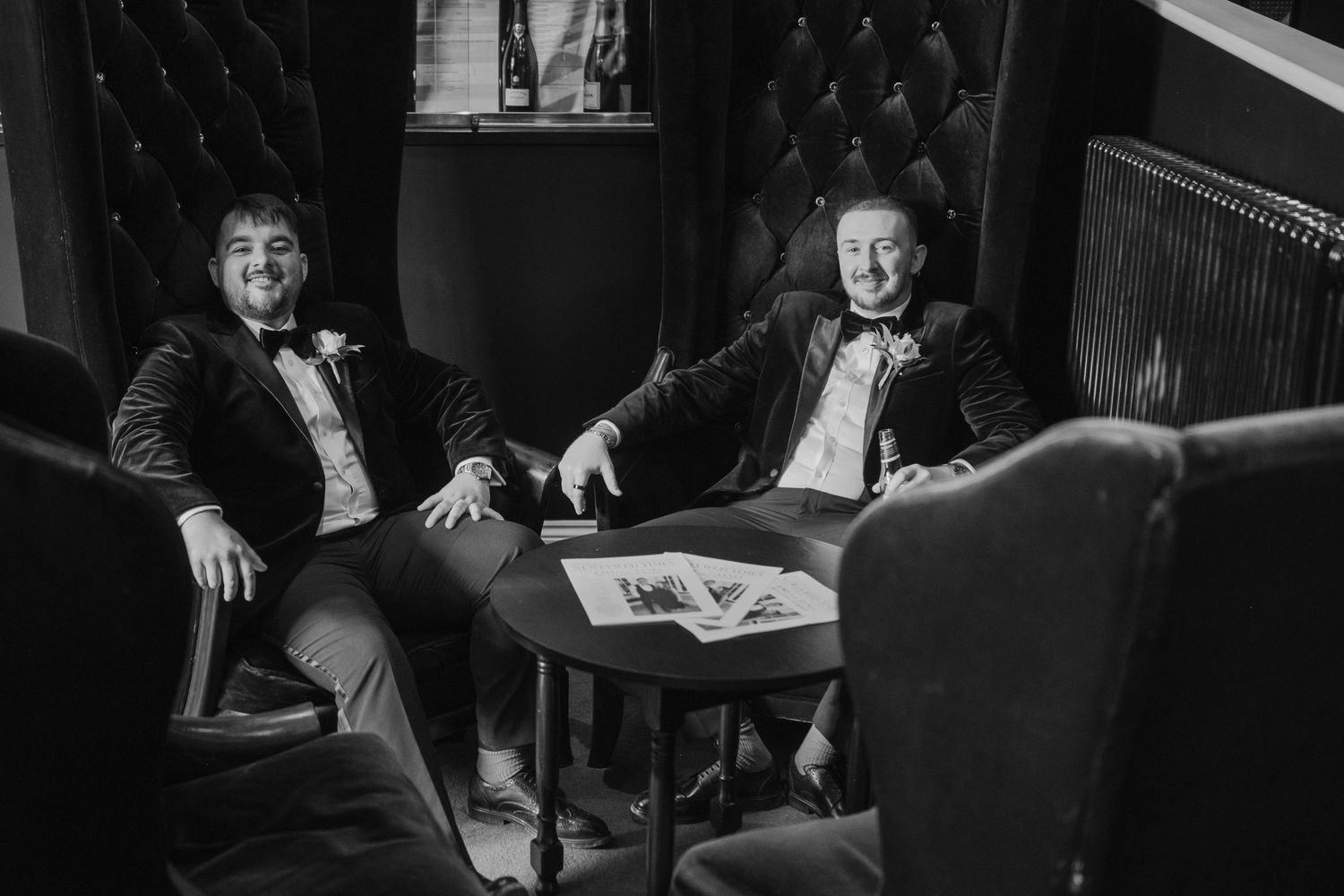 Two men in tuxedos with bow ties relaxing in velvet chairs at a small table in a dimly lit lounge.