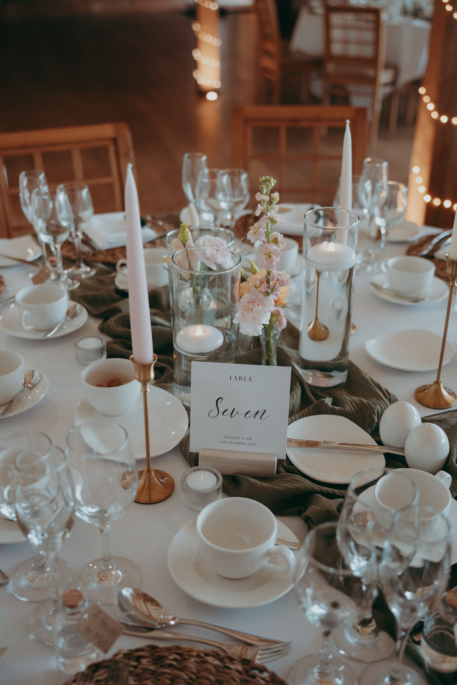 Elegant wedding reception table setting with white china, gold candlesticks, blush florals, and a Table Seven place card.