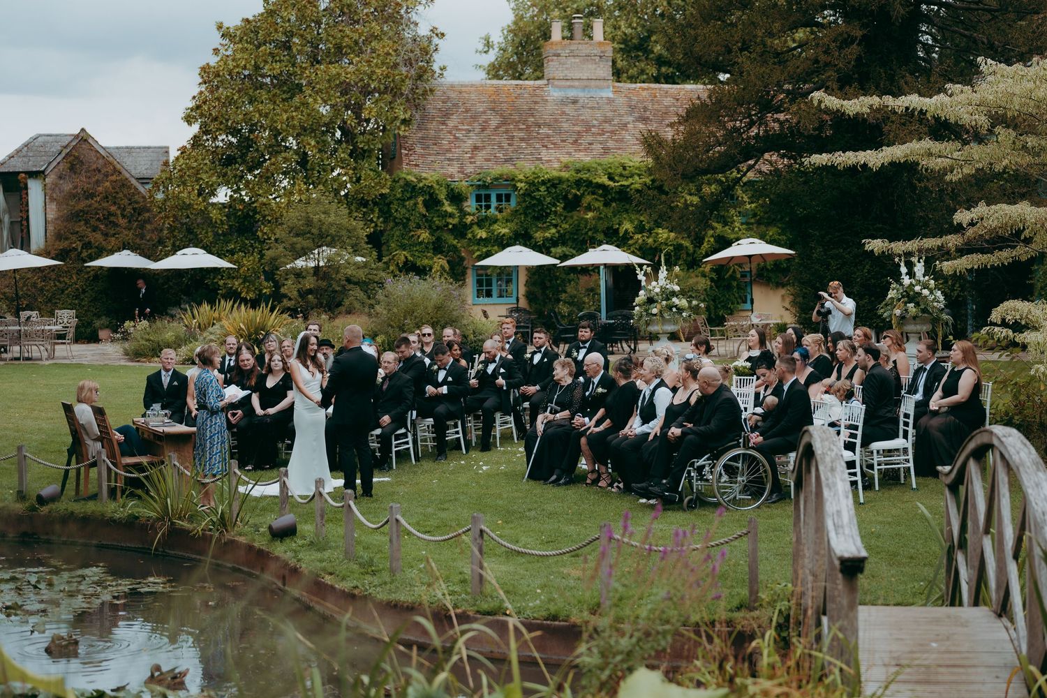 Outdoor wedding ceremony in a garden setting with guests seated on chairs near a pond, brick building backdrop.
