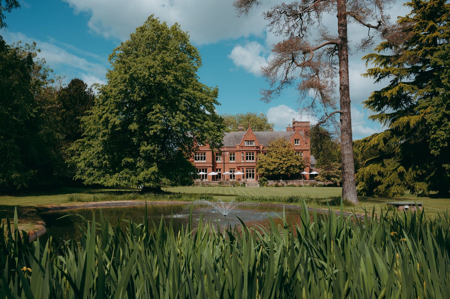 Red brick English manor house reflected in a tranquil pond, surrounded by lush green trees and gardens on a sunny day.