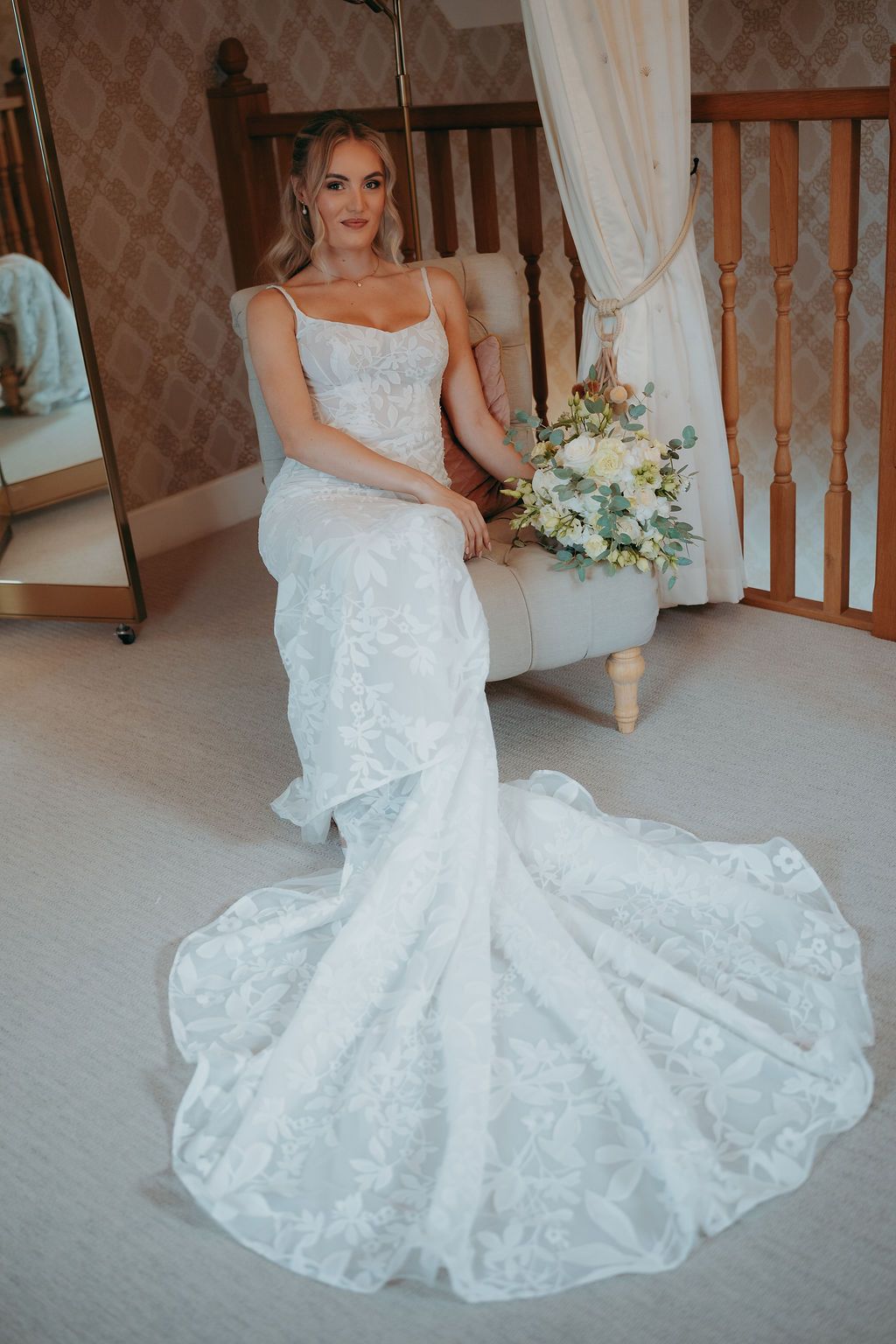 Bride in a white lace mermaid wedding gown with a long train sits on a cream armchair holding a pastel bouquet of white and green flowers.