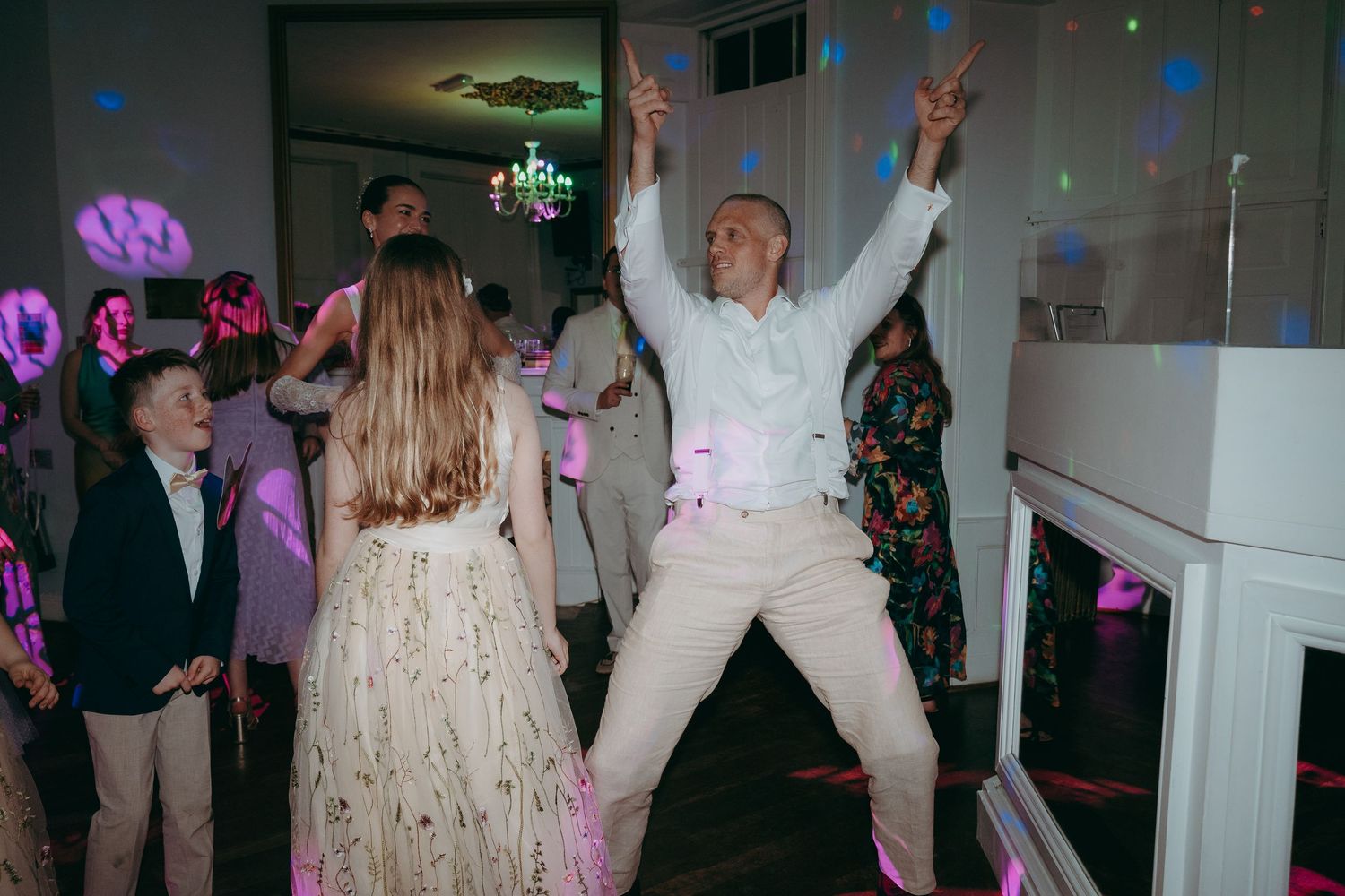 Man in white suit dancing energetically at a wedding reception with guests celebrating on the dance floor.