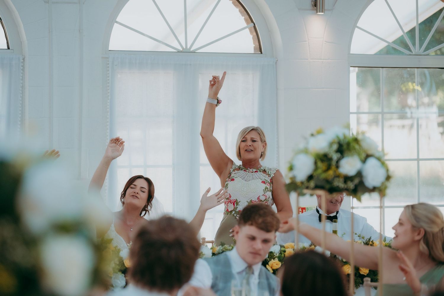 Joyful wedding reception guests celebrating with raised arms in an elegant white venue with floral arrangements.