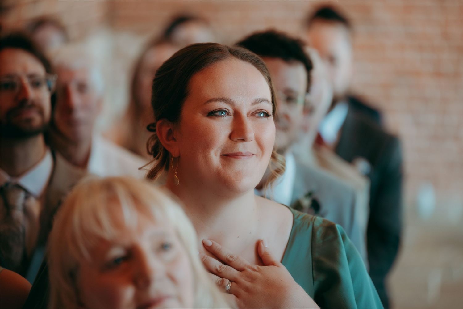 Emotional wedding guest in teal dress smiling with hand on chest during ceremony.