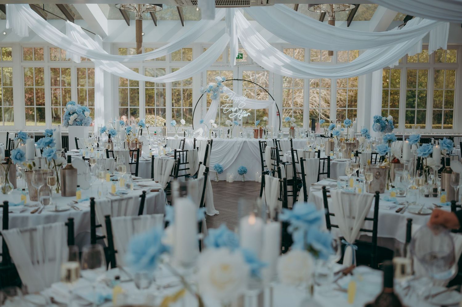 Elegant wedding reception in a greenhouse venue with white draping, blue floral centerpieces, and round tables set for guests.