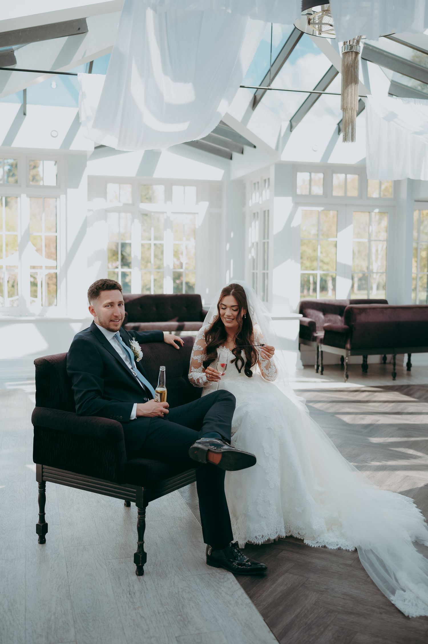 Bride and groom relaxing in elegant conservatory venue, bride in lace gown, groom in navy suit, holding champagne.