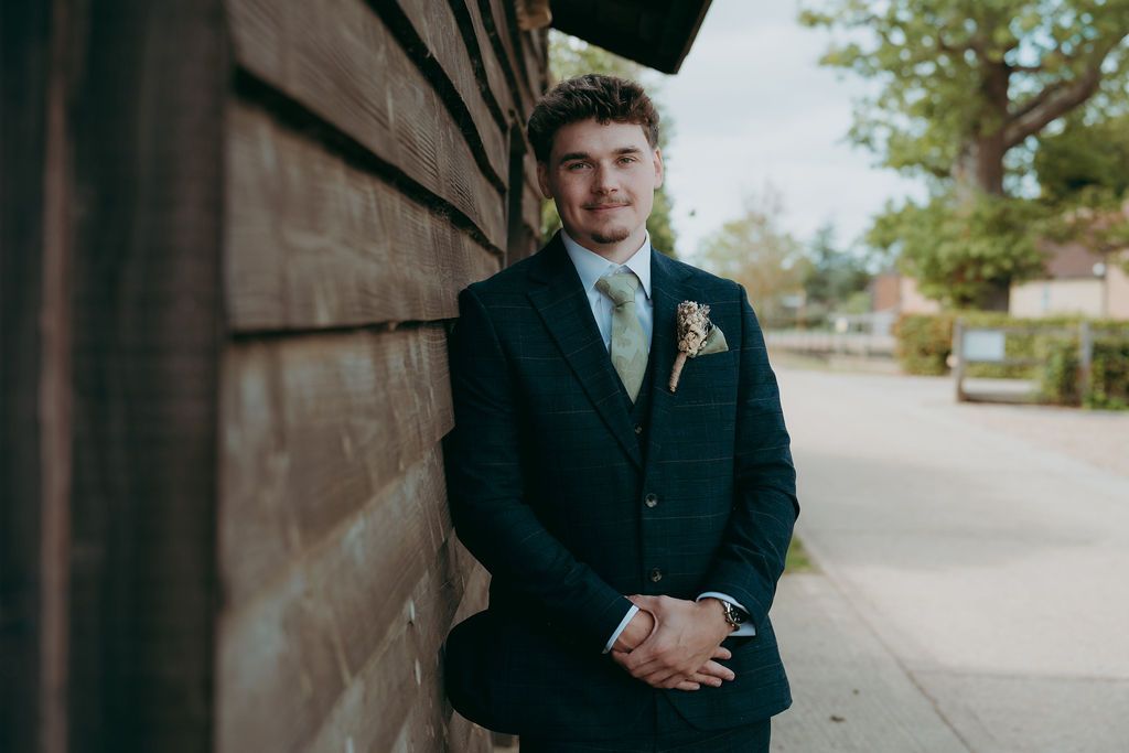 Smiling young man in a navy suit with a floral boutonniere stands with hands clasped beside a rustic wooden wall on a quiet street.