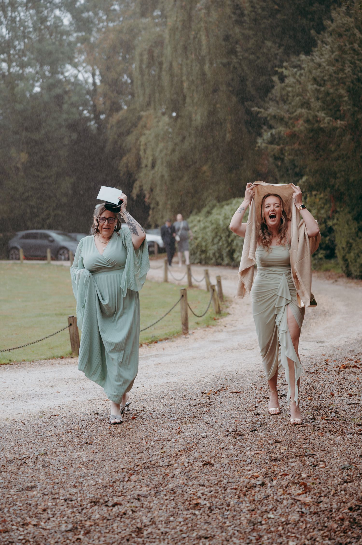 Two women in sage green dresses laugh while covering their heads walking along a gravel path at an outdoor wedding.