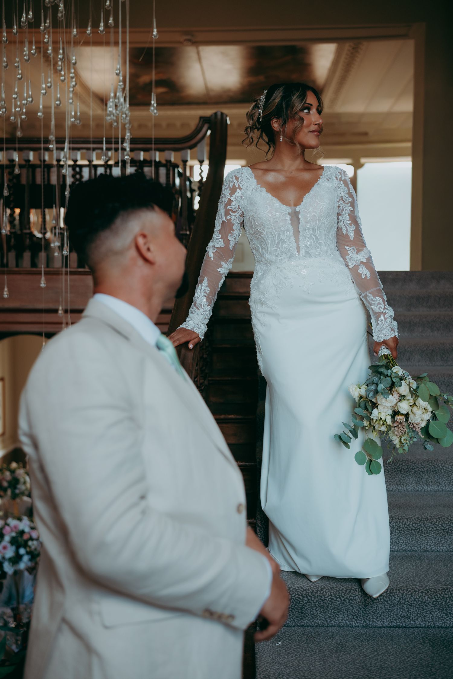 Bride in lace long-sleeve wedding gown holding bouquet on staircase with groom in white suit in foreground.