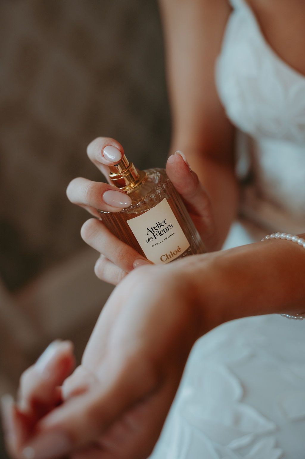 A bride in a white wedding dress sprays Chloé Atelier des Fleurs perfume on her wrist while getting ready.