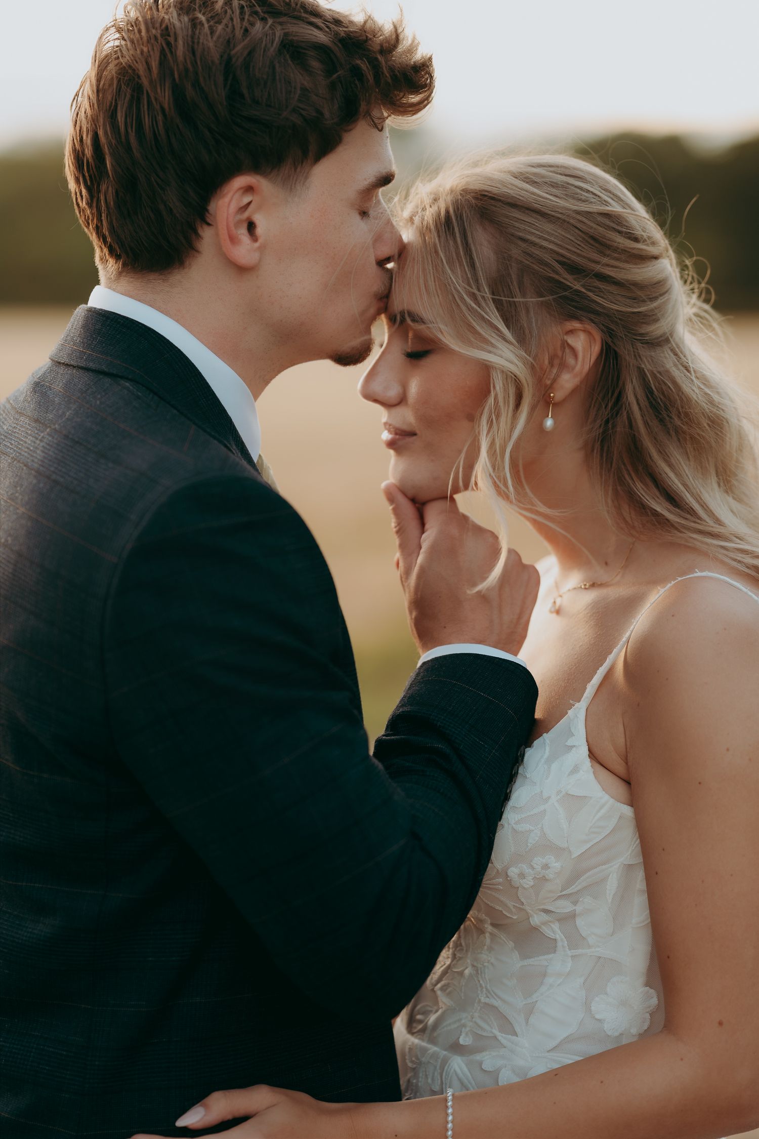 A groom in a dark suit tenderly kisses his bride on the forehead as she smiles in a white lace wedding dress.
