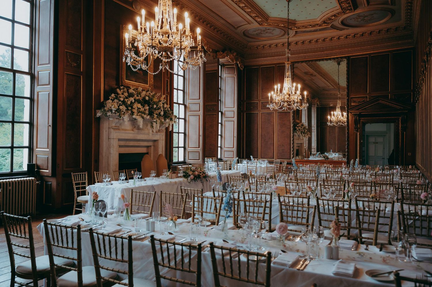 Elegant wedding reception in a grand ballroom with crystal chandeliers, wood paneling, and long tables with floral centerpieces.