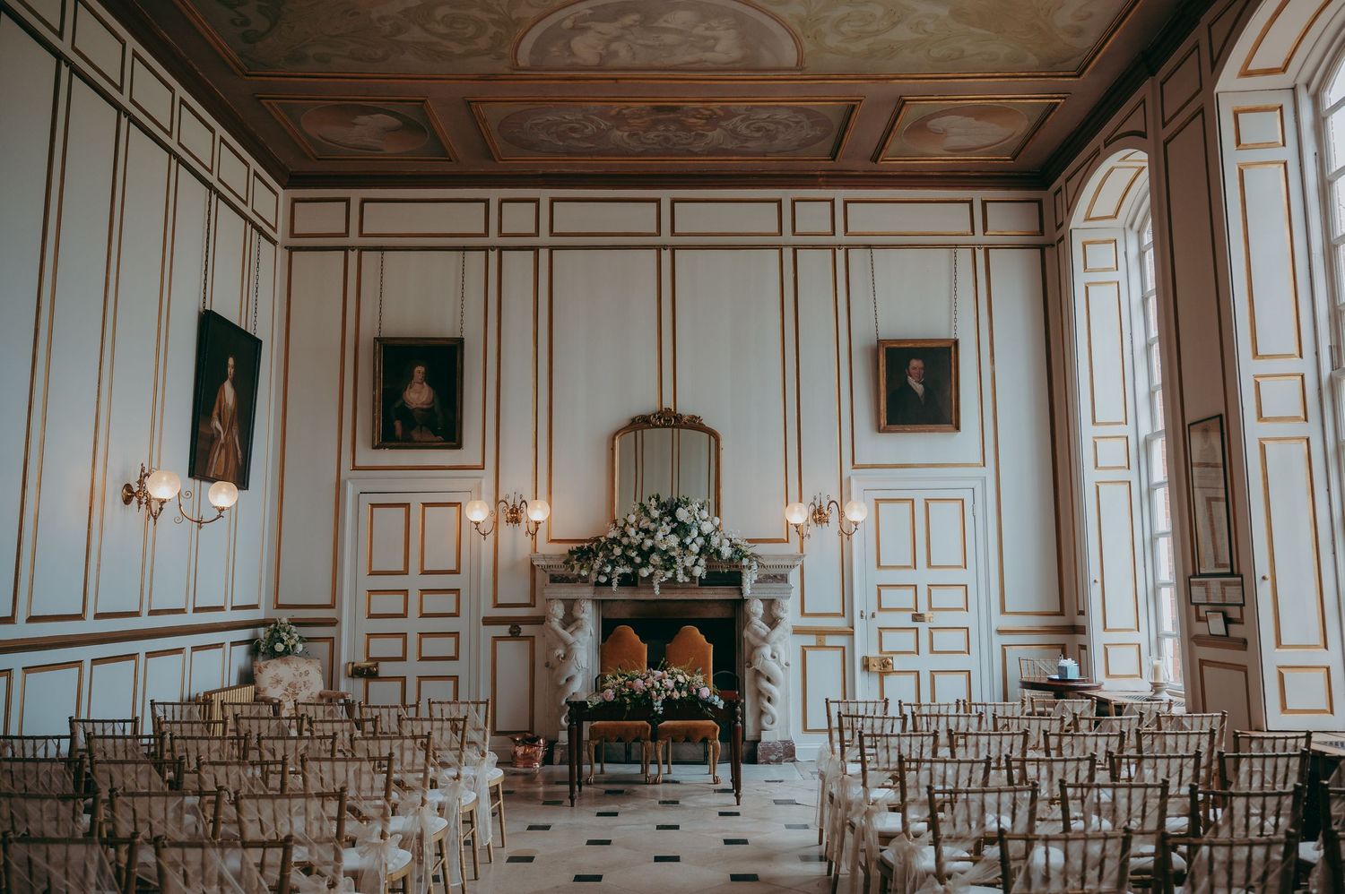 Elegant wedding ceremony room with gold-trimmed white paneling, portrait paintings, chiavari chairs, and floral fireplace display.