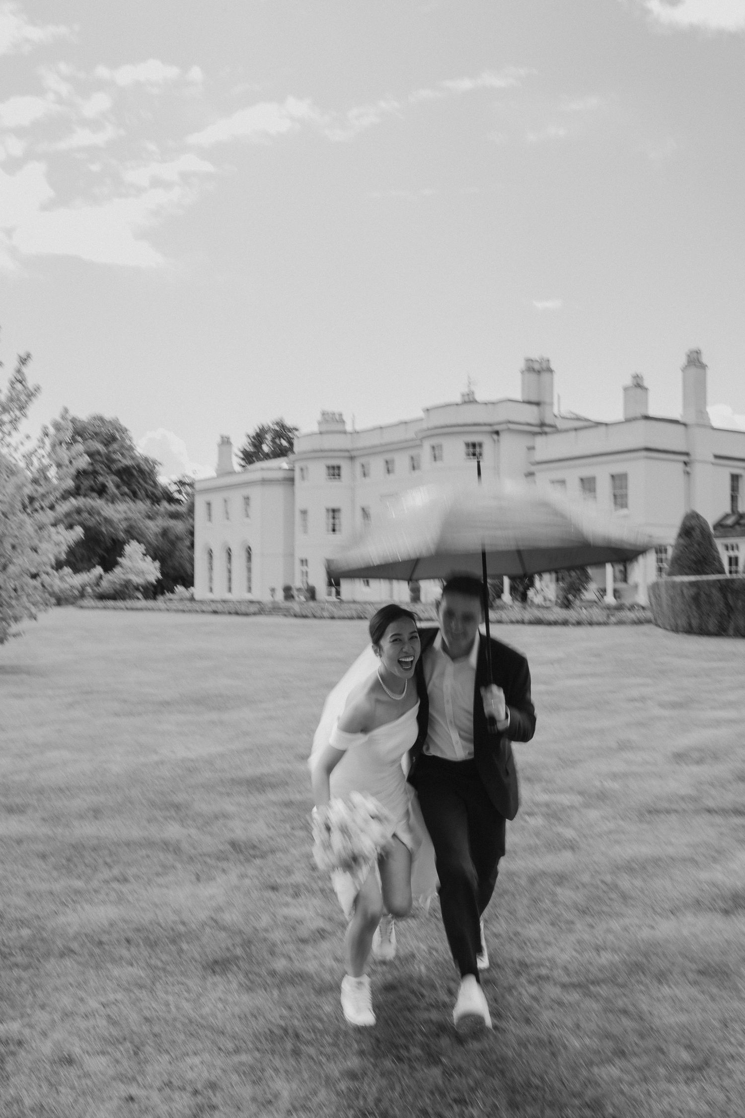 A joyful couple runs across a lawn sharing an umbrella outside a grand estate on their wedding day.
