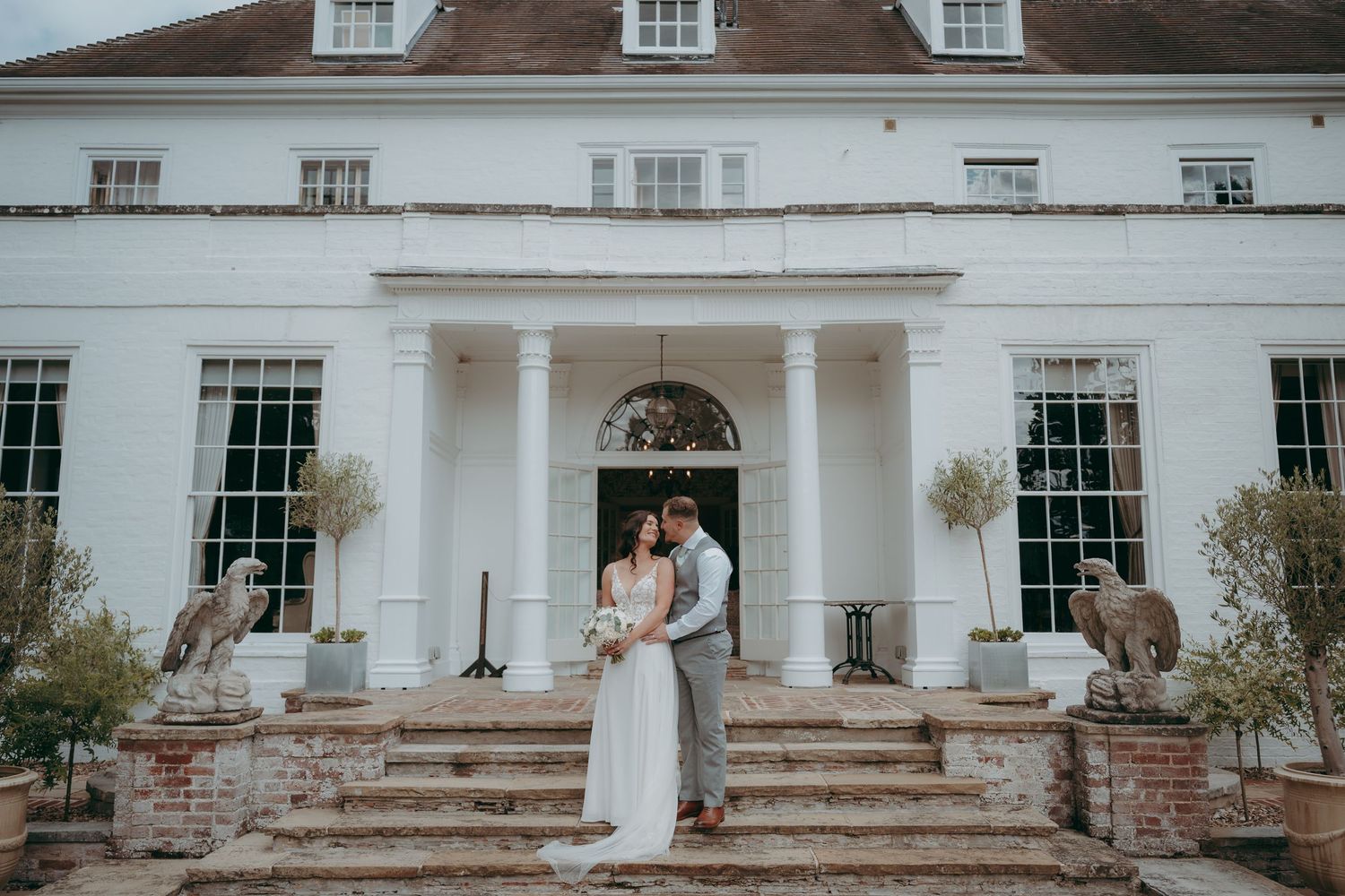 Bride and groom embrace on brick steps of elegant white Georgian manor house wedding venue with columned entrance.