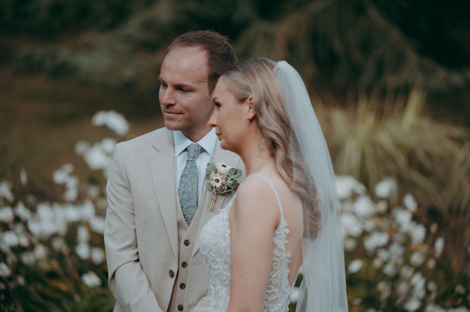 Bride and groom in elegant wedding attire standing among white wildflowers, sharing a tender moment outdoors.