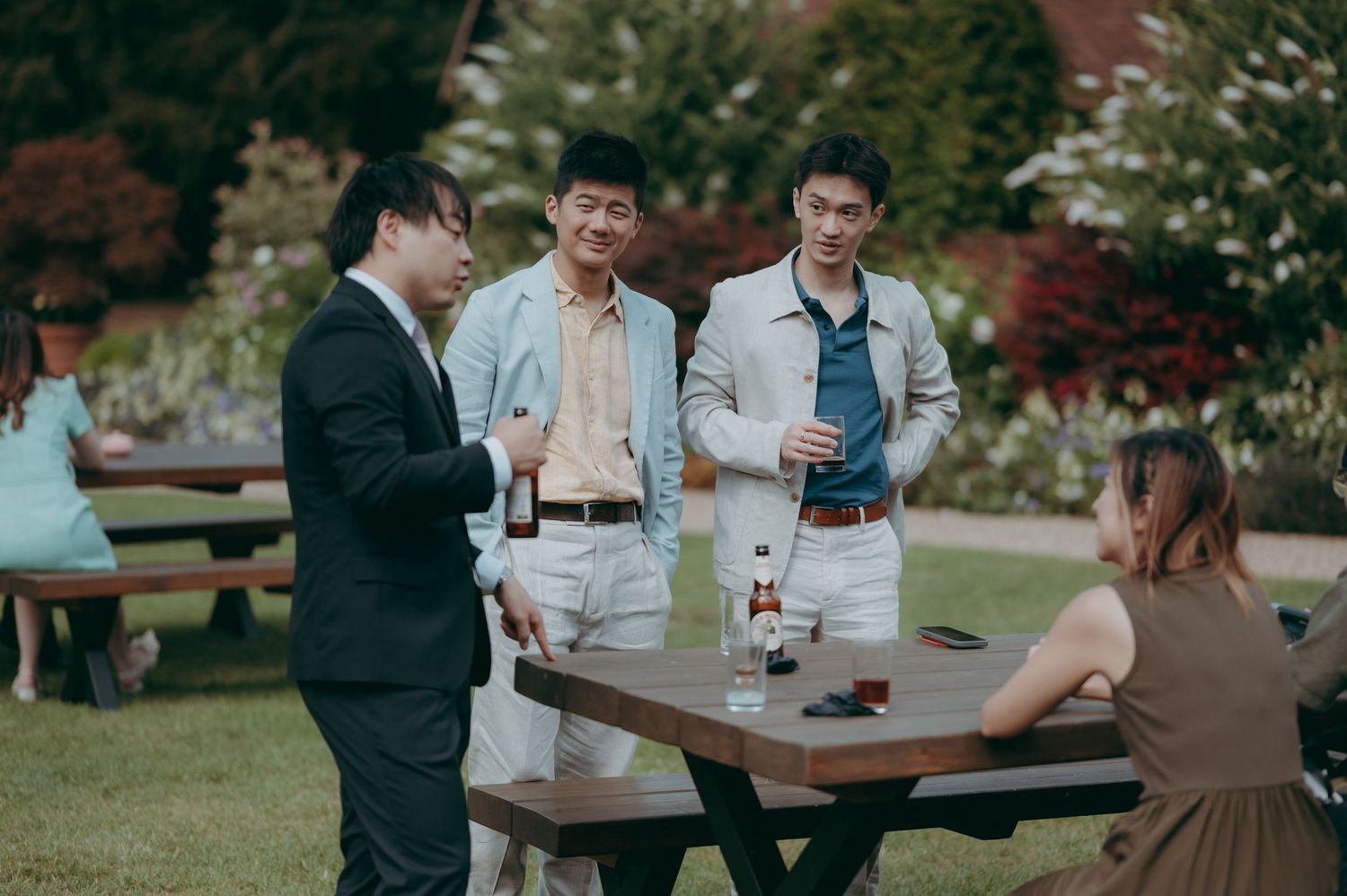 Three young men socializing outdoors at a picnic table with drinks, dressed in smart casual attire in a garden setting.