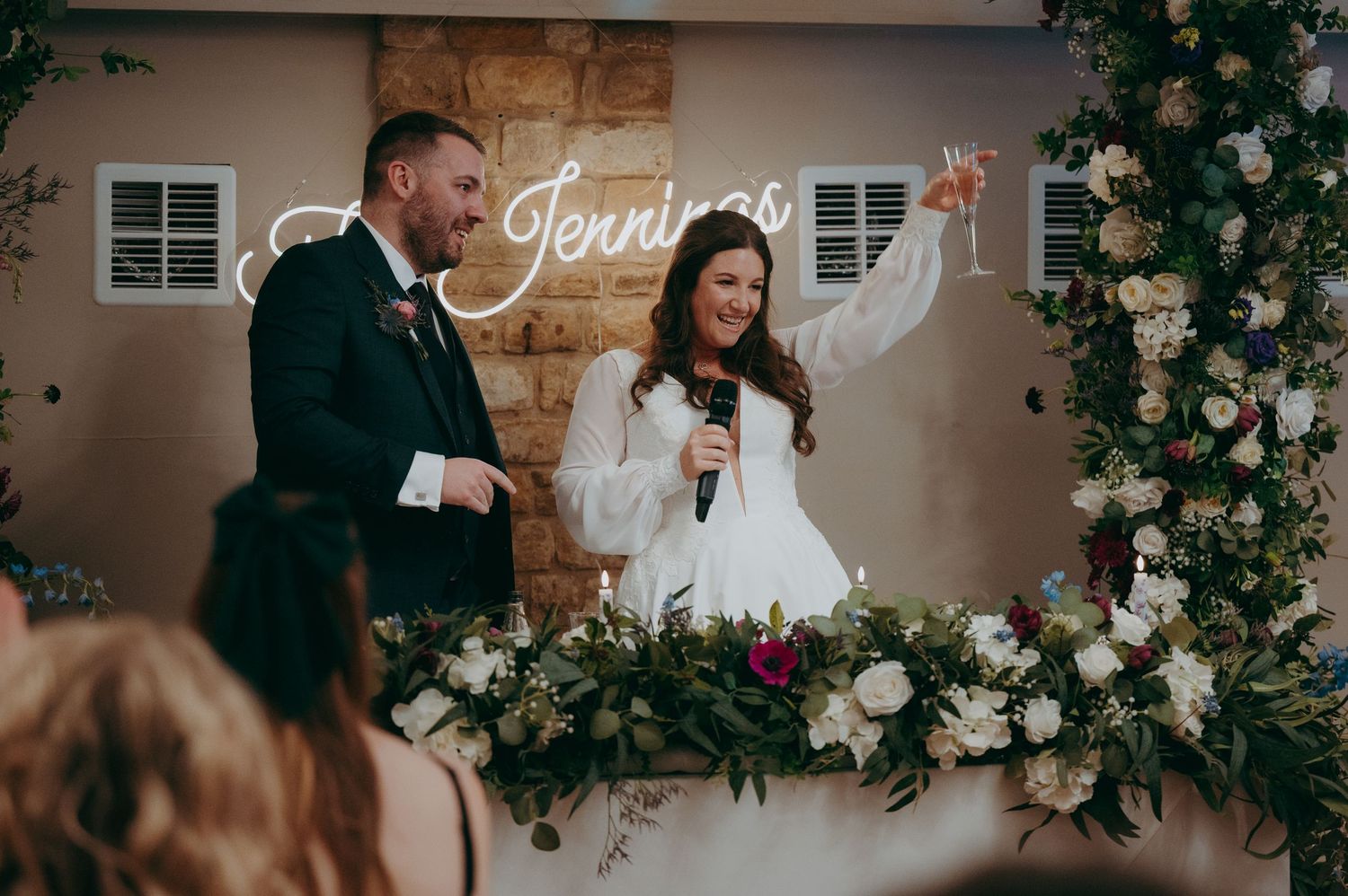 Bride and groom toast at wedding reception with floral arch and neon sign backdrop.