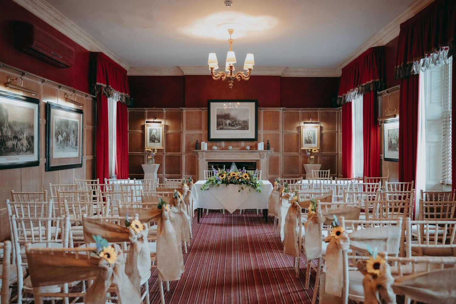 Elegant wedding ceremony room with chiavari chairs, floral arrangements, fireplace, and red drapes in a classic venue.