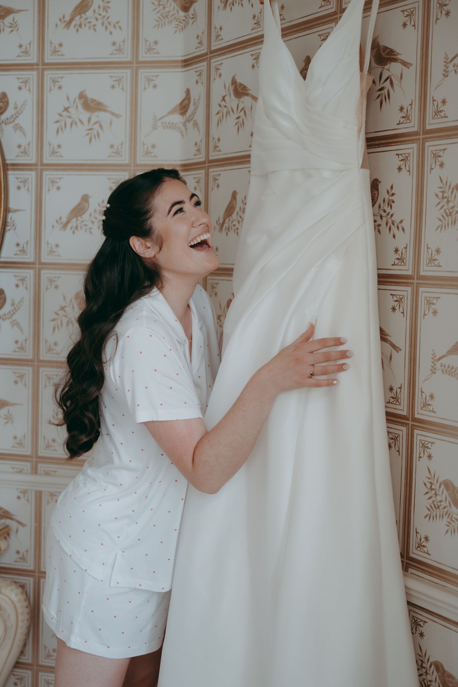 Joyful bride in white pajamas laughing while embracing her wedding dress hanging on a floral wallpapered wall.