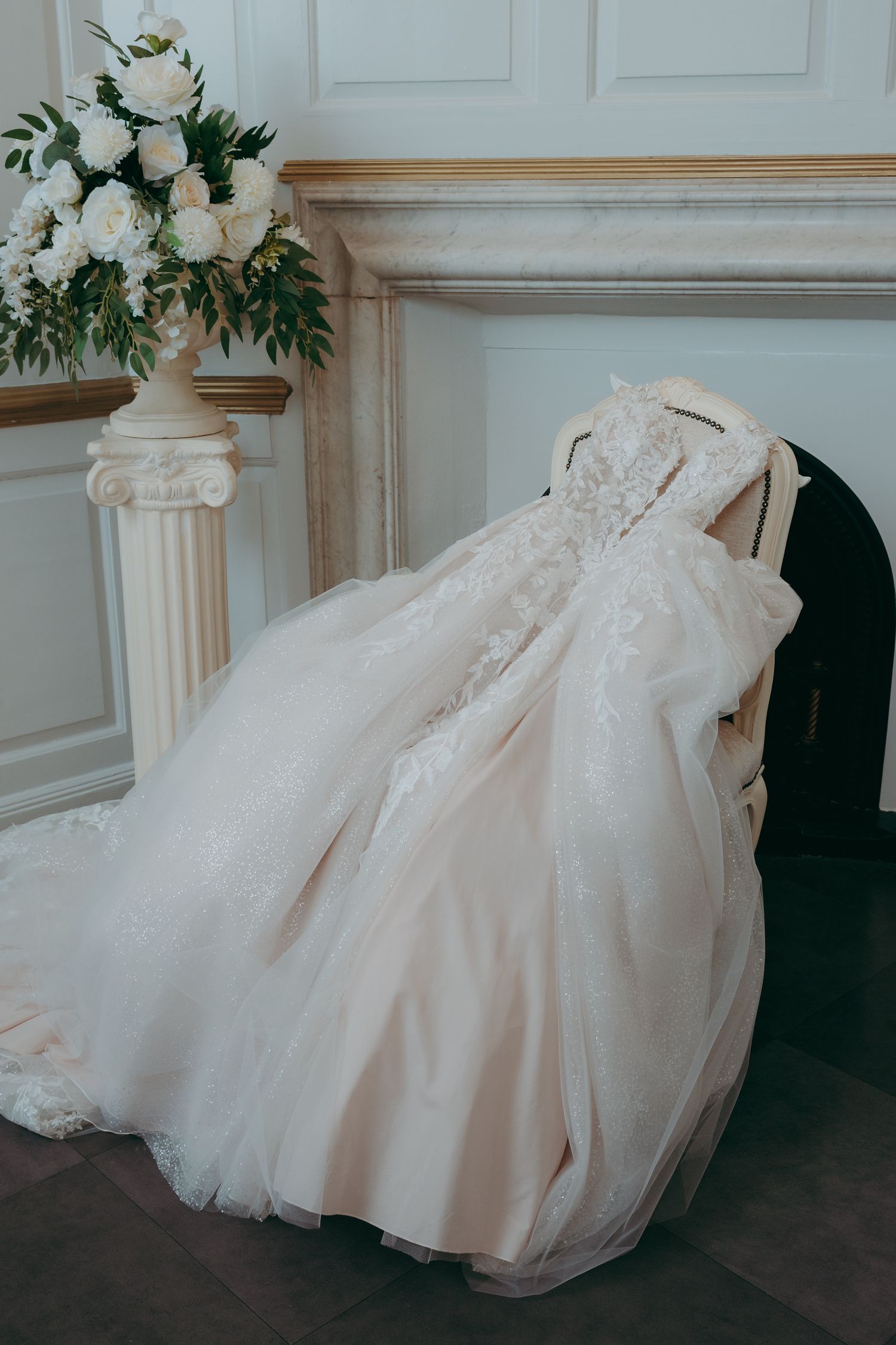 Elegant blush wedding gown with lace detailing draped over a chair near a fireplace and white floral arrangement.