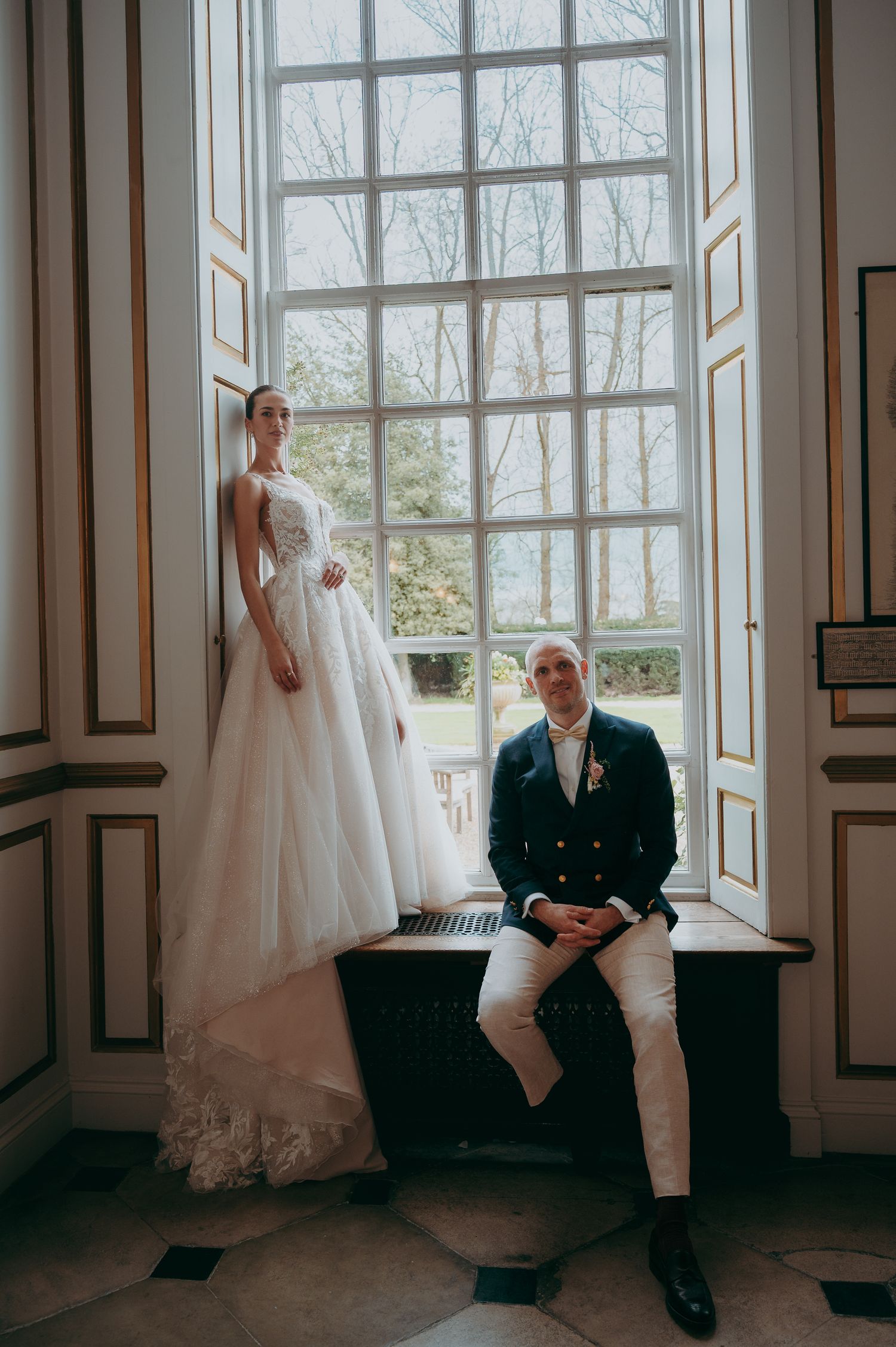 Bride in white ball gown stands beside groom in navy suit on window seat in elegant gold-trimmed room.