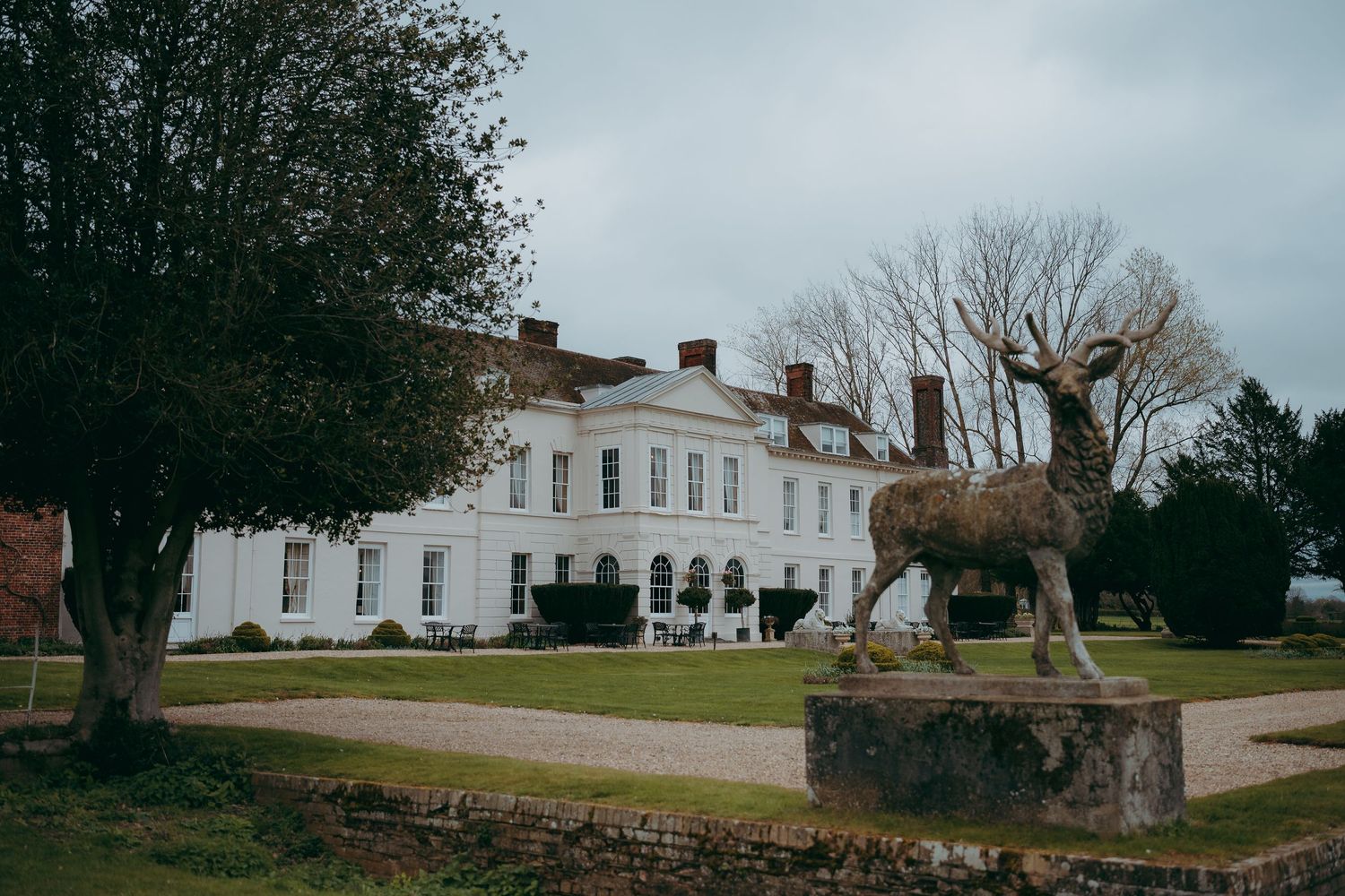 A topiary deer statue stands before a grand white Georgian country house with manicured grounds on an overcast day.