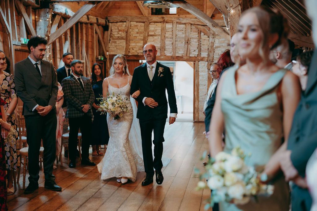 Bride in a lace gown holding a bouquet walks down the aisle with an older man in a rustic barn wedding venue as guests look on.