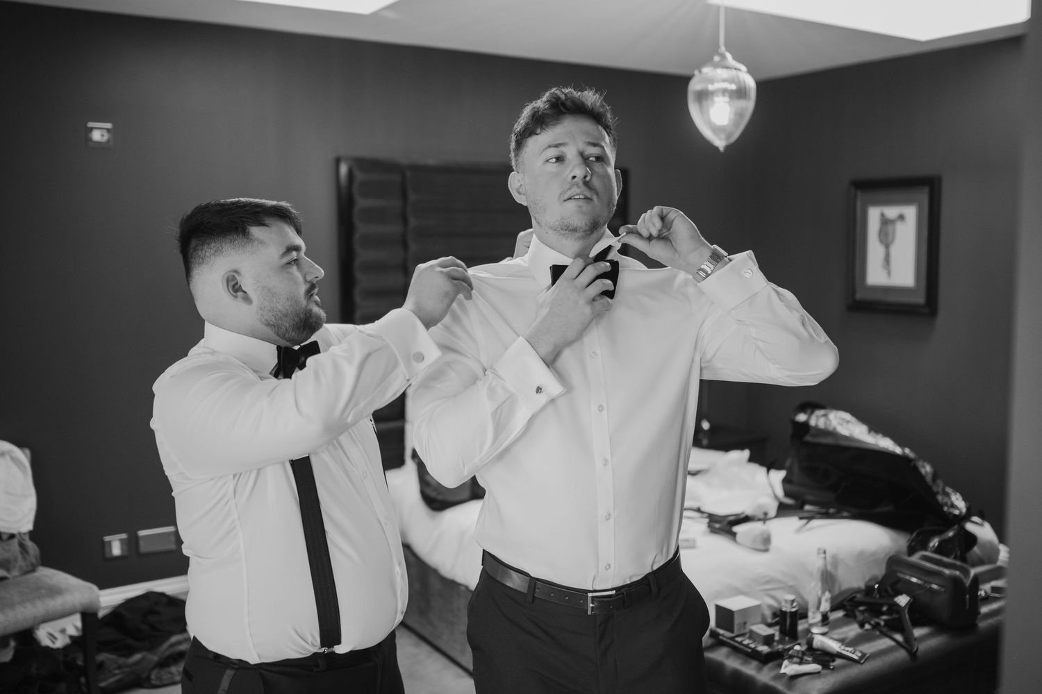 Two men in formal white dress shirts adjusting ties and collar in black and white wedding preparation photo.