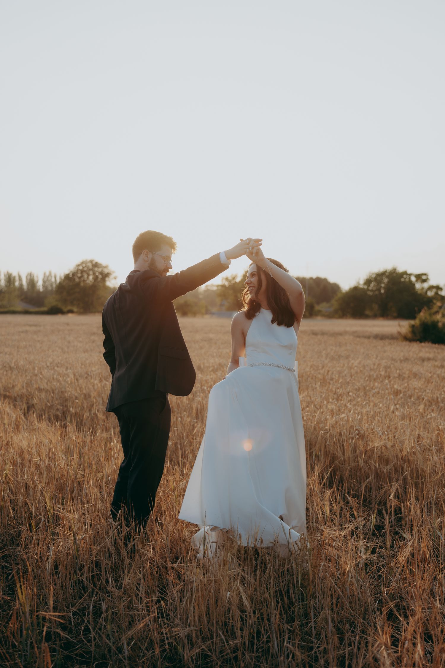 Bride in off-shoulder white ball gown and groom in black tuxedo posing on a tree-lined gravel path.
