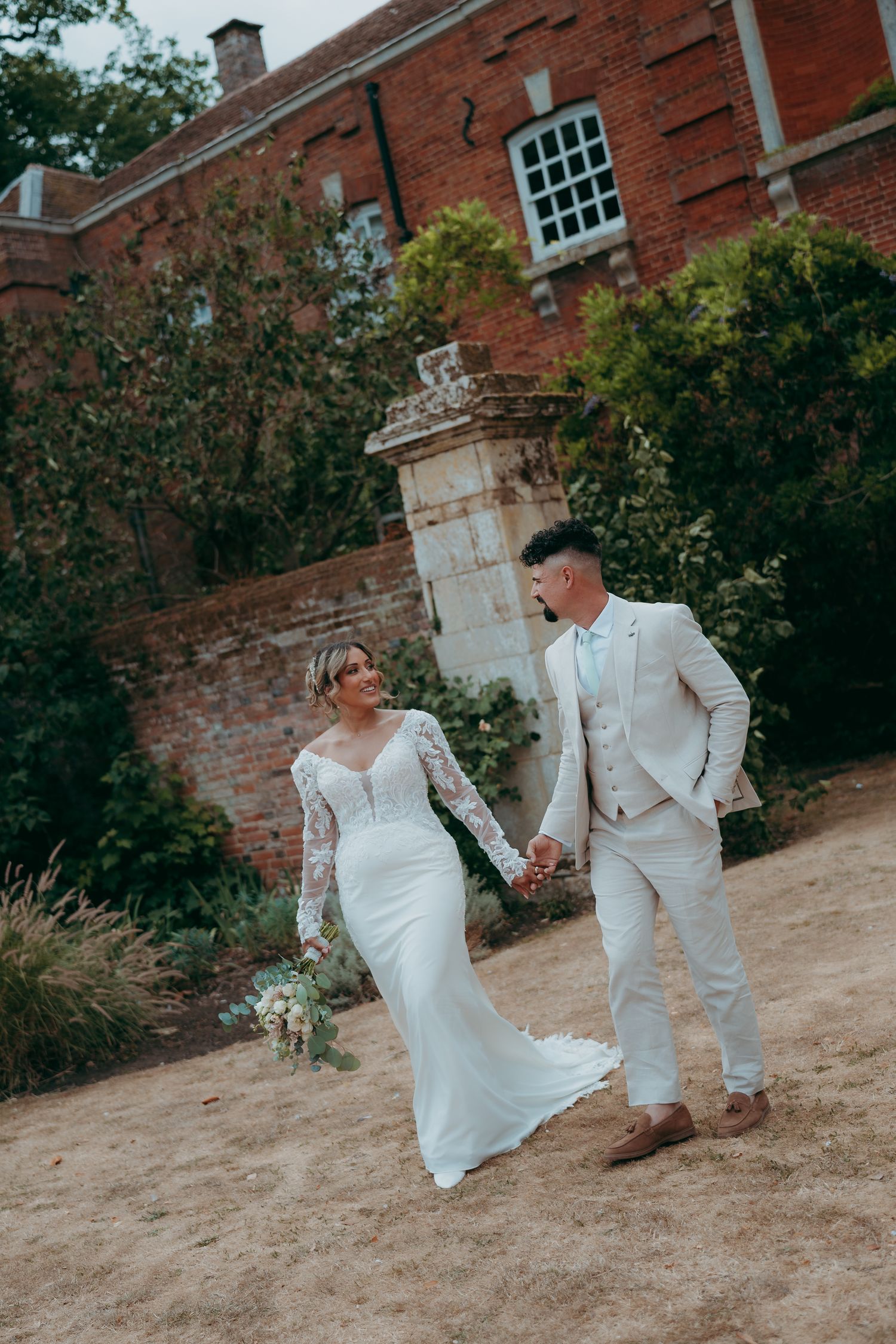 Bride and groom holding hands walking in front of a brick manor house, bride in lace gown, groom in light suit.