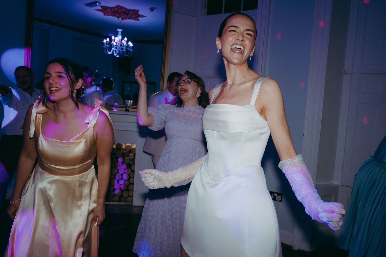 Joyful bride in white dress and gloves dancing at wedding reception with guests in colorful evening wear.