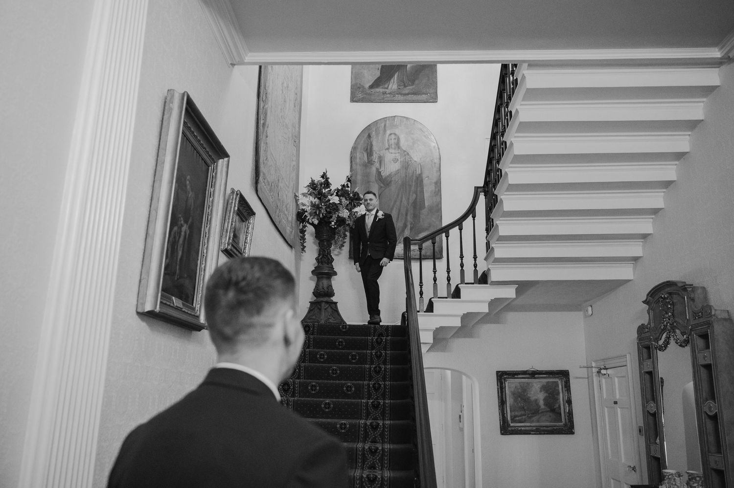 Black and white wedding photo of bride and groom descending an elegant staircase with floral arrangement.