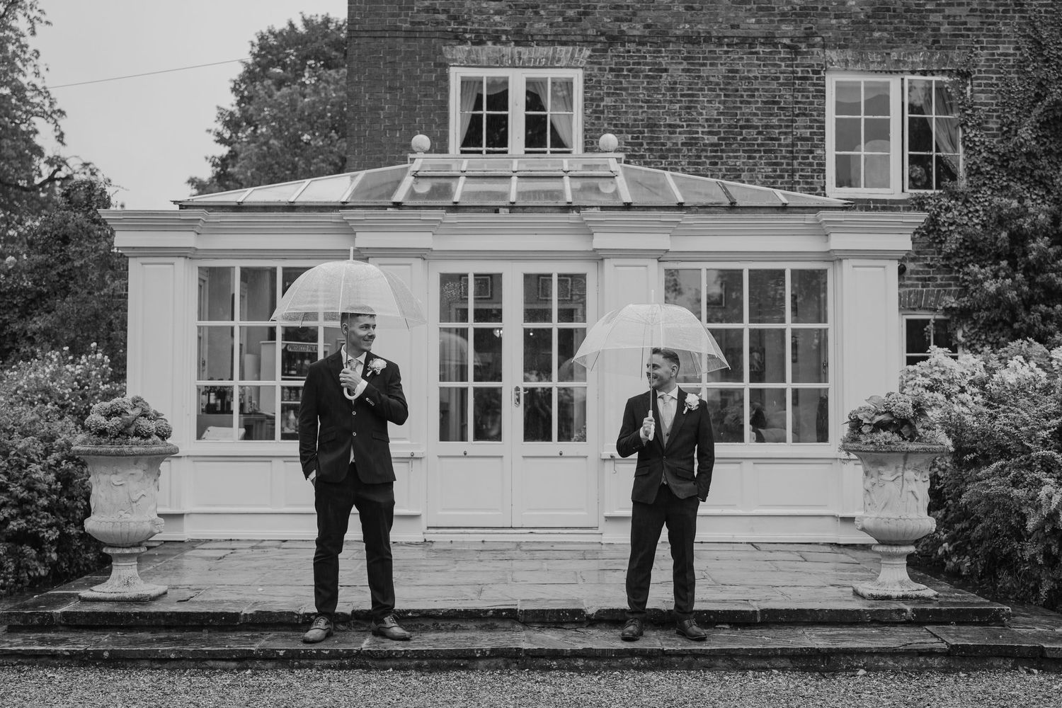 Two grooms in suits holding clear umbrellas stand before an elegant manor house in this black and white wedding photo.