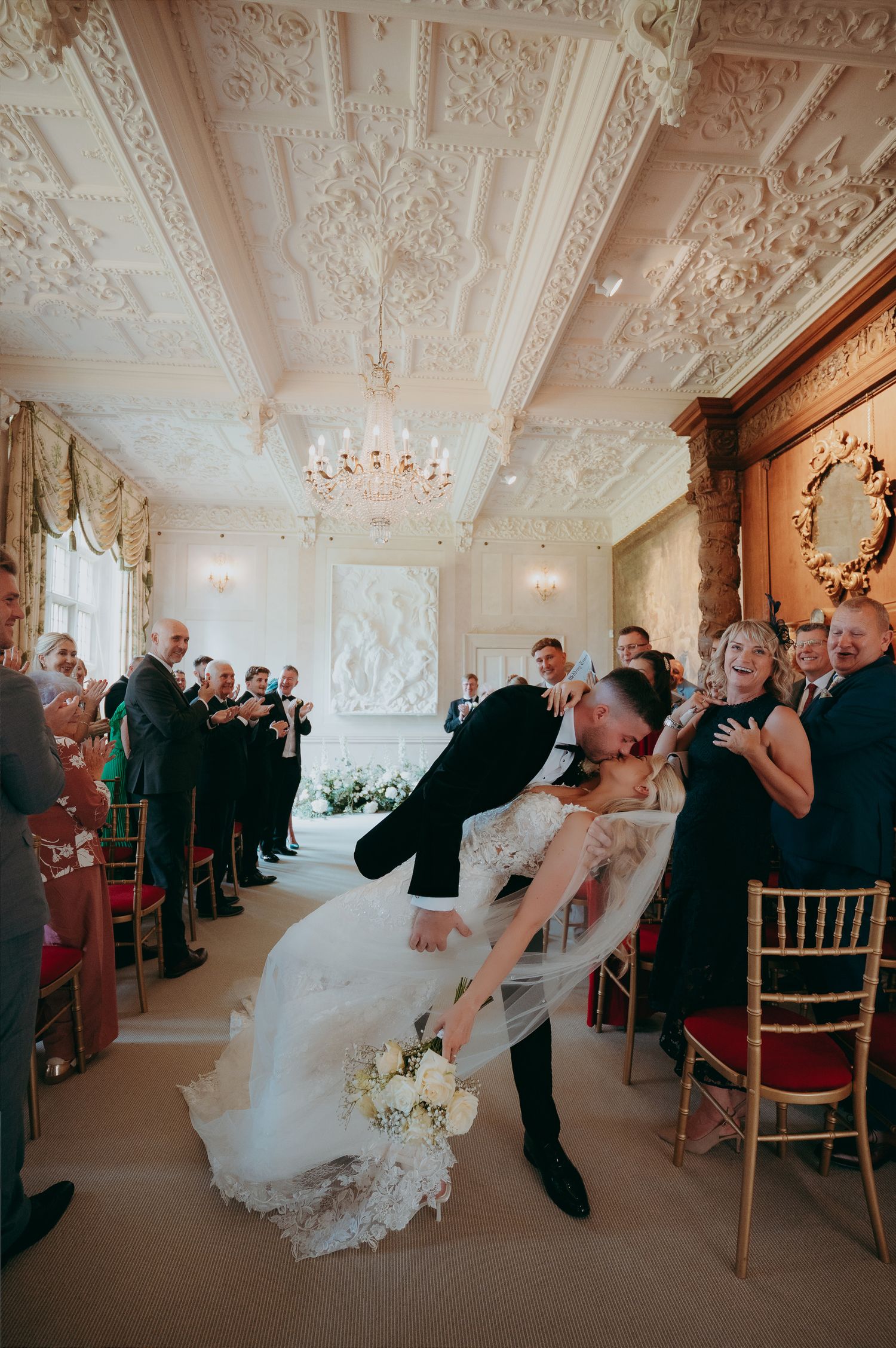 Bride in white off-shoulder gown and groom in black tuxedo hold champagne glasses in rustic brick venue.