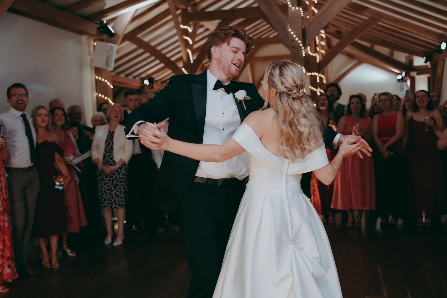 Bride and groom share first dance at rustic barn wedding reception, guests watching in background.